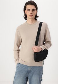 Beige neulepaita, jossa on pyöreä pääntie, yhdistettynä vaaleansinsisiin farkkuihin. Musta suorakulmion muotoinen olkalaukku, jossa on vetoketju ja olkahihna.