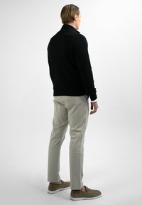 Man staat met zijn rug naar de kijker, gekleed in een zwart kabelbreisel trui, lichtbeige broek en olijfgroene instappers met witte zolen.