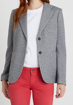 Frau trägt einen grauen Blazer über einem weißen Hemd, kombiniert mit roten Hosen, steht vor einem schlichten hellen Hintergrund.
