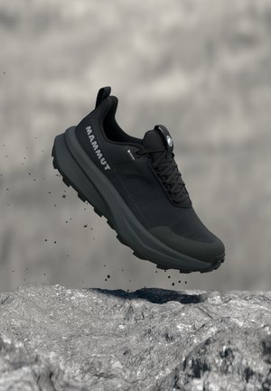 Chaussure de randonnée noire Mammut avec une semelle épaisse flottant au-dessus d'un sol rocheux, montrant des particules de terre tombant de la semelle.