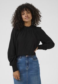 Black long-sleeve blouse with a high neckline, smocked detailing, and fitted cuffs, paired with a blue denim skirt.