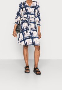 Femme portant une robe portefeuille à motifs longueur genou avec des carrés bleus et beiges, des sandales noires, et un sac à bandoulière noir.