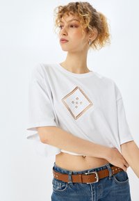 Hvid cropped T-shirt med et kvadratisk broderet design med blomster mønstre, parret med blå denim jeans og et brunt bælte med studs.