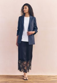 Granatowa marynarka w prążki na biały top, zestawiona z czarną spódnicą z koronkowym wykończeniem i czarnymi sandałami na paski na drewnianej podłodze.