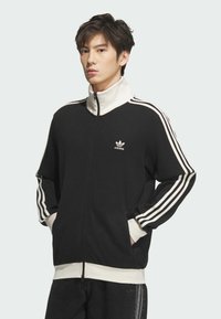 Crna rebrasta jakna na zatvarač s visokim ovratnikom, s bijelim trakama na rukavima i izvezenim adidas logotipom na prsima.