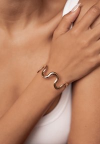 Bransoletka w odcieniu złota z falistym wzorem, gładką teksturą i otwartym stylem mankietu, noszona na skórze o tonie ciała z białą bluzką.