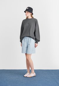Oversized grå sweatshirt, lyseblå teksturerte shorts, svart bøttehatt og rosa sko med hvite stropper; står på et blått gulv.