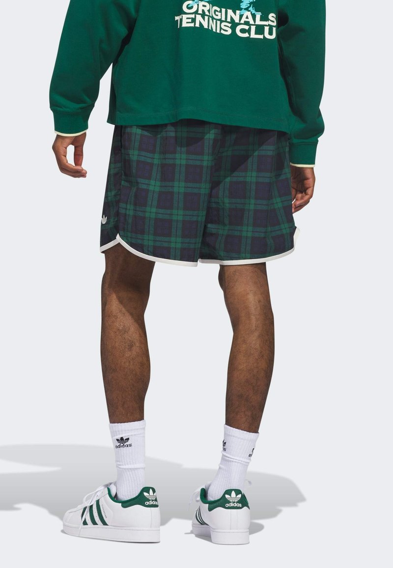 Grünes langärmliges Oberteil, karierte Shorts in Grün und Marineblau, weiße Besätze, sportliche weiße Schuhe mit grünen Akzenten und Logo, weiße Knöchelsocken.