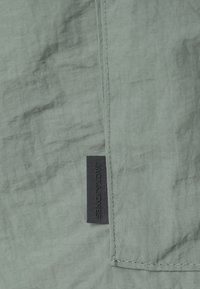 Lichtgroene stof met een gekreukt textuur, met een zwart rechthoekig label met de tekst "JACK & JONES" op de zijkant geborduurd.