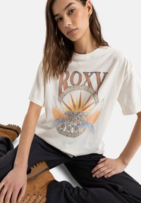 Kremowa koszulka z nadrukiem "ROXY" w kolorze pomarańczowym, z grafiką słońca i desek surfingowych, w połączeniu z ciemnymi spodniami i brązowymi butami.