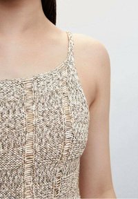 Haut en maille présentant un mélange de couleurs beige et blanc, avec des bretelles fines et des motifs texturés, mettant en valeur un design en maille ouverte.