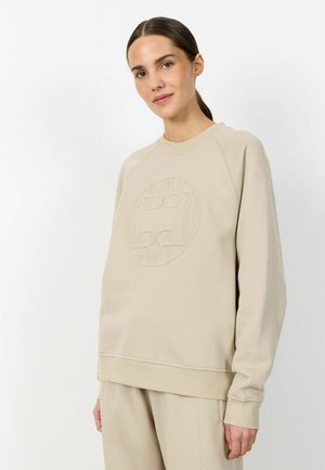 Mlada ženska v bež usklajeni jopa in hlačah z izbočenim okroglim logotipom, stoji pred belim ozadjem.