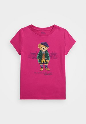 Jasnoróżowy t-shirt z krótkim rękawem z misiem w zielonej kurtce, niebieskim berecie i paskowanych skarpetkach na tle szkicu budynku.