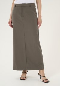 Jupe maxi en tissu marron foncé avec une texture lisse et un détail de fente à l'avant. Portée avec des sandales à talons hauts noires à lanières.