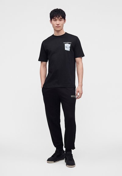Νέος άνδρας στέκεται φορώντας μαύρο T-shirt με λογότυπο στην τσέπη του στήθους και μαύρο παντελόνι jogger με αθλητικά παπούτσια σε απλό φόντο.