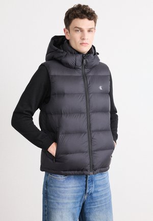 Zwarte puffer vest met een capuchon, voorzien van een ritssluiting, zijzakken en een gewatteerd ontwerp. Draagt over een zwarte langemouwenshirt.