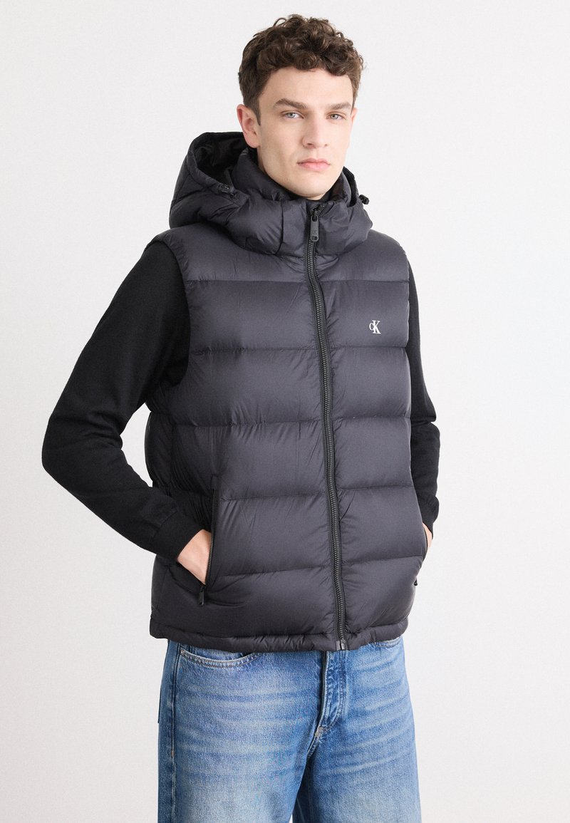 Zwarte puffer vest met een capuchon, voorzien van een ritssluiting, zijzakken en een gewatteerd ontwerp. Draagt over een zwarte langemouwenshirt.