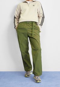 Ljusbeige zip-up-tröja med svarta ränder och logotyp, kombinerad med gröna byxor med elastisk midja, bälte och två sidofickor.