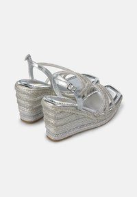 Sandalias de cuña plateadas con tacón de textura trenzada, tirante delgado al tobillo y diseño de puntera abierta. Presenta acentos metálicos y un acabado suave.