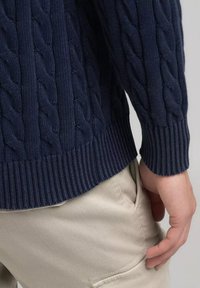 Donkerblauwe kabelgebreide trui met geribbelde zoom en manchetten, gemaakt van gestructureerde stof; hand van het model op een beige broekzak geplaatst.