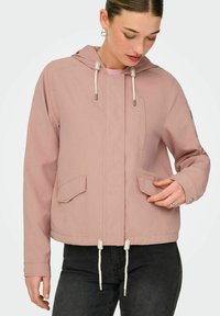 Femme portant une veste à capuche rose poussiére avec des poches avant et des cordons, associée à un pantalon noir, regardant vers le bas et ajustant sa manche.