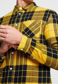 Koszula w żółto-czarną kratę na guziki, z kieszenią na piersi, wykonana z lekko teksturowanej tkaniny z białymi guzikami.
