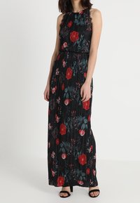 Robe maxi noire plissée avec un motif floral rouge et vert. Design à encolure halter, taille cintrée, sans manches, portée avec des sandales à talons noires.