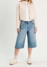 Veste courte beige en tissu texturé, associée à des culottes en denim bleu clair et des baskets blanches. Design simple avec des détails pratiques.