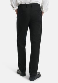 Pantaloni neri con vestibilità dritta, dotati di due tasche posteriori con chiusura a bottoni e una texture liscia, abbinati a scarpe nere.