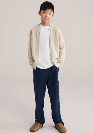 Niño joven de pie con las manos en los bolsillos, vistiendo un suéter beige con cremallera, camiseta blanca, pantalones azul marino y zapatos marrones sin cordones, fondo neutral.