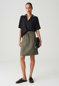 Woman wearing a black short-sleeve top with white trim, olive drawstring skirt, black slip-on shoes, holding a black bag.