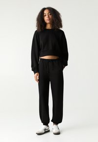Pulover scurt negru dintr-un material moale, asortat cu pantaloni jogger negri, cu talie elastică și manșete. Se poartă adidași albi.