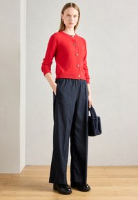 Červený pletený cardigan s kulatým výstřihem a zlatými knoflíky, kombinovaný s tmavě pruhovanými kalhotami s širokými nohavicemi a malou strukturální námořnickou kabelkou.