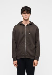Sweat à capuche zippé marron en mélange de coton avec poche avant, capuche à cordon de serrage et poignets côtelés, assorti à un pantalon foncé.