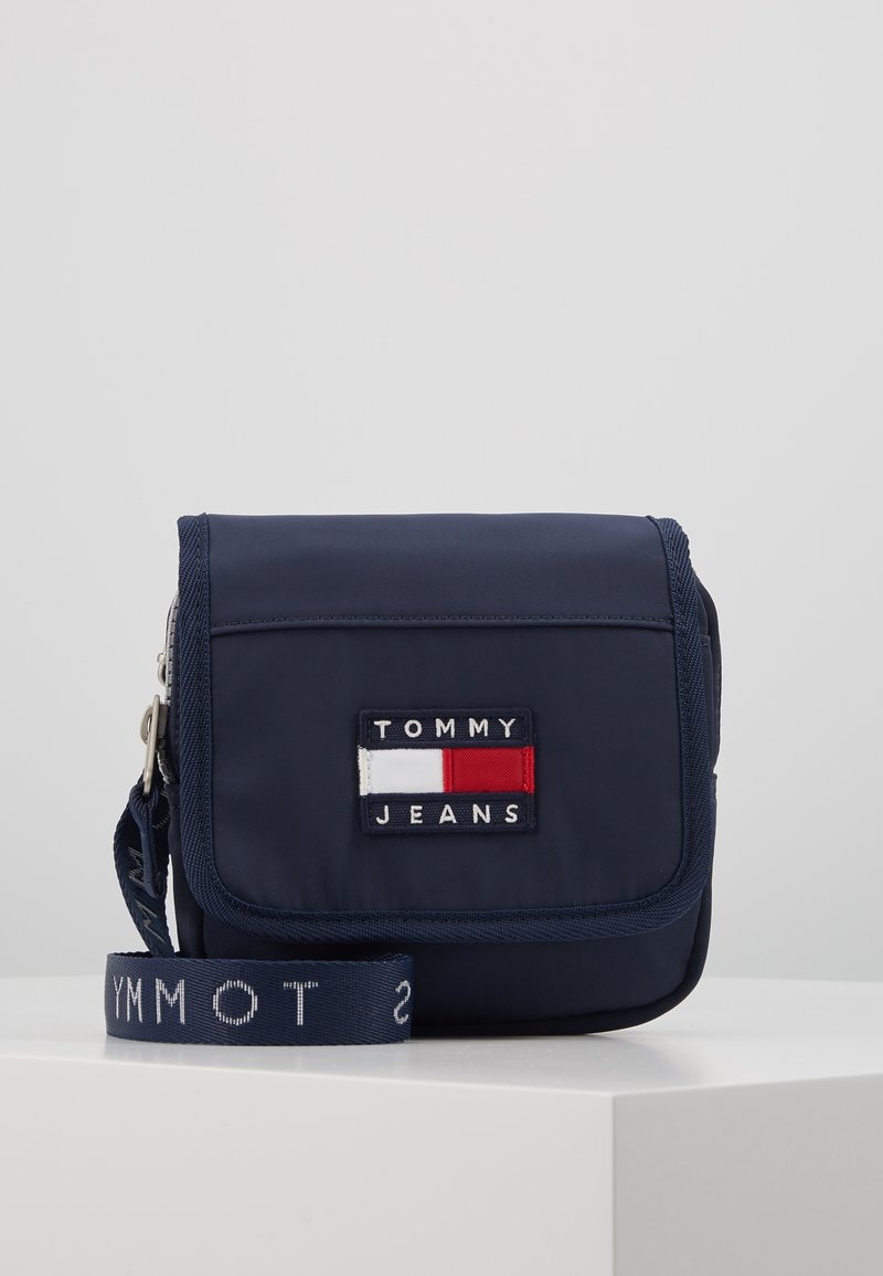 Borsa a tracolla in tessuto blu navy con pattina anteriore, caratterizzata da un patch "TOMMY JEANS" e una tracolla intrecciata con dettagli testuali. Design compatto e funzionale.