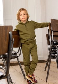 Olijfgroene sweatshirt met capuchon en knopen, gecombineerd met bijpassende sweatpants met gestructureerde patches. Casual sneakers in rood en geel.