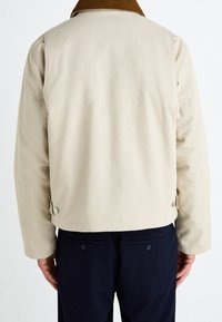 Chaqueta de color crema con un cuello marrón acanalado, con un corte recto, botones en los laterales y una textura suave. Usada sobre pantalones oscuros.