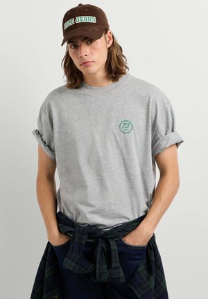 Jonge man met een bruine pet die "Pepe Jeans," leest, draagt een grijs T-shirt met opgerolde mouwen en een donker geruit overhemd om zijn middel geknoopt.