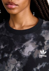 Mustavalkoinen ja harmaa collegepaita, jossa on maastokuvio, resoroitu pyöreä pääntie sekä valkoinen Adidas-logo rinnassa. Pehmeä, teksturoitu kangas.