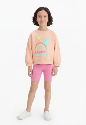 Copil în picioare, purtând un hanorac piersică cu soare, curcubeu, palmier și textul „Happy Summer”, pantaloni scurți roz și pantofi albi slip-on, pe fundal alb.