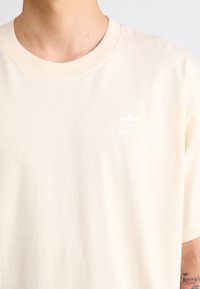 Světlý broskvový tričko s krátkým rukávem vyrobené z bavlny, s bílým logem Adidas na levé části hrudi. Hladká textura a design s kulatým výstřihem.