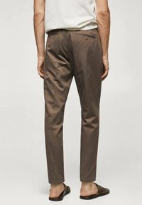 Pantalon marron à coupe ajustée, avec poches arrière, coutures discrètes et texture lisse. Associé à des sandales foncées.