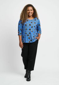 Zils rakstains tops ar ģeometriskiem dizainiem un melniem akcentiem, saskaņots ar melniem biksēm un potīšu zābakiem. Mīksta auduma un relaksēta piegriezuma.