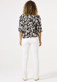 Bloes met bloemenpatroon in crème en zwart, voorzien van opgerolde mouwen en een geraffineerde achterkant, gecombineerd met strakke witte jeans en beige schoenen.