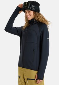 Black textured jacket with a high collar, zippered pockets, and thumbholes, paired with yellow ski pants and a black knit beanie.