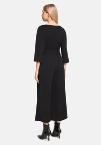 Czarny kombinezon z długimi rękawami, dopasowaną talią, szerokimi nogawkami i suwakiem z tyłu. Noszony z czarnymi botkami za kostkę.