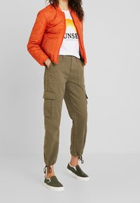 Femme portant une veste matelassée orange, un t-shirt blanc avec un motif arc-en-ciel, un pantalon cargo vert olive et des baskets sans lacets.