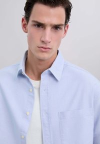 Camicia button-up a righe azzurre, con taschino, realizzata in cotone. Presenta un colletto classico e una t-shirt bianca sotto.