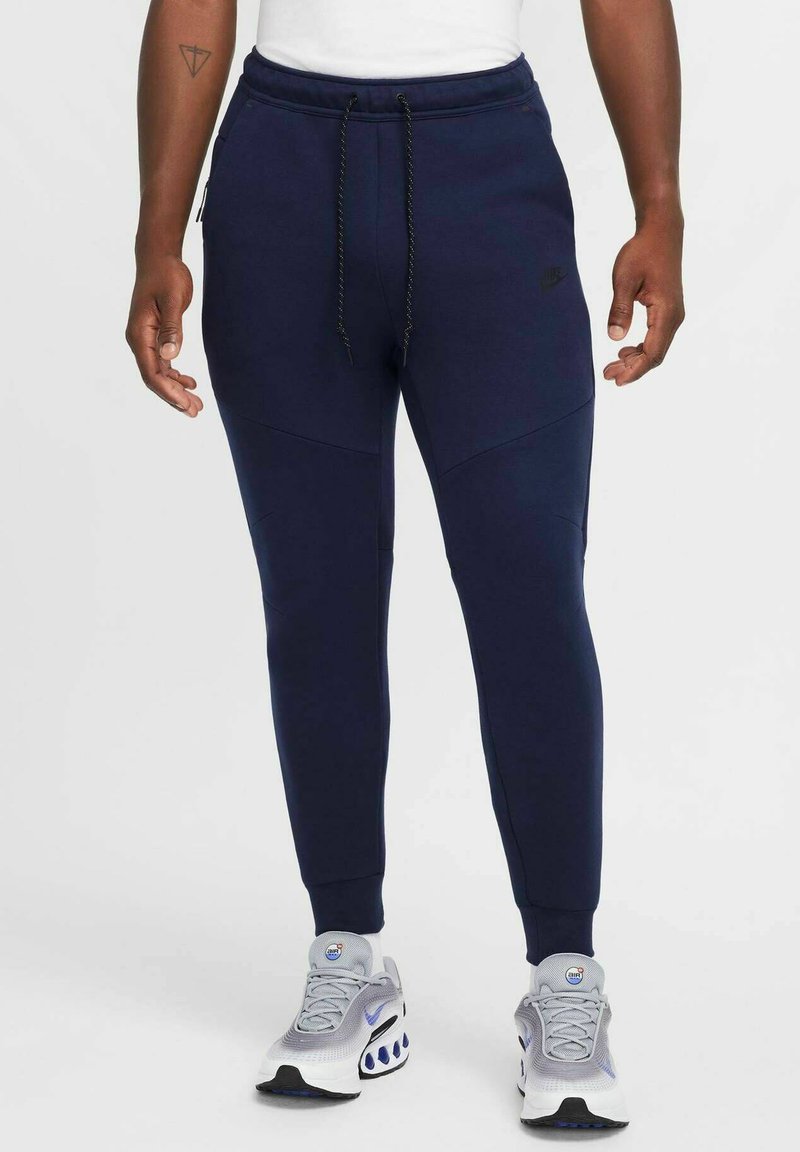 Marineblå Nike sweatpants med en tætsiddende pasform, elastisk talje og snøre. Har sidelommer og teksturerede paneler. Stylings med grå sneakers.
