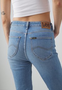 Blå denimjeans med hög midja, har en läderetikett på baksidan samt två bakfickor med böjda sömmar och kontrasterande sömnad.